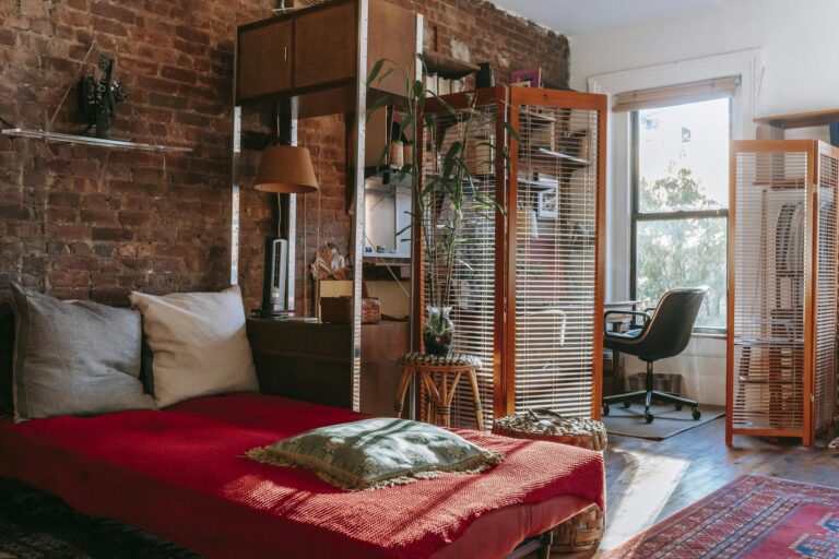 Comfortable bed with red cover and pillows placed near brick wall in cozy room with isolated workspace decorated with houseplants near window